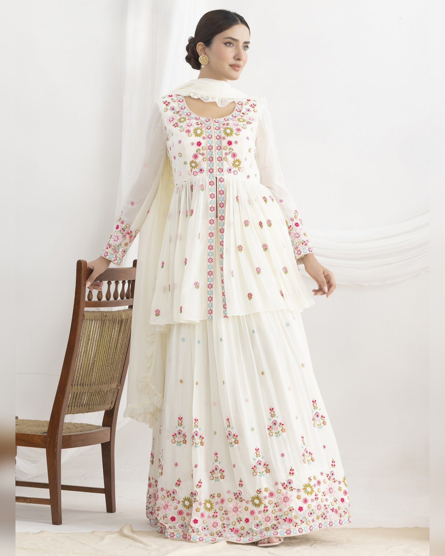 Woman wearing a white floral dress standing in a room with a wooden chair and white curtain.