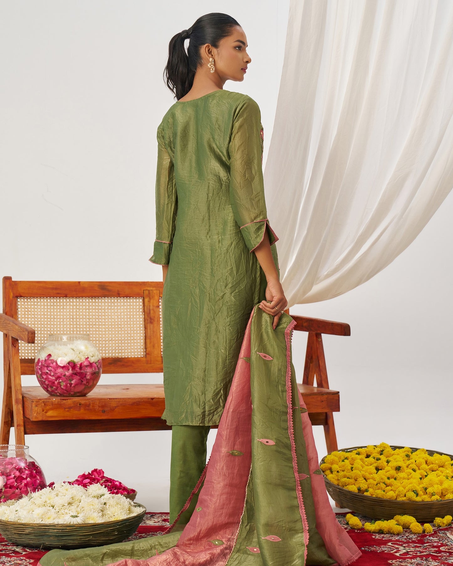 Woman in a green traditional outfit with a pink dupatta, standing in a decorated room with flowers and a chair.