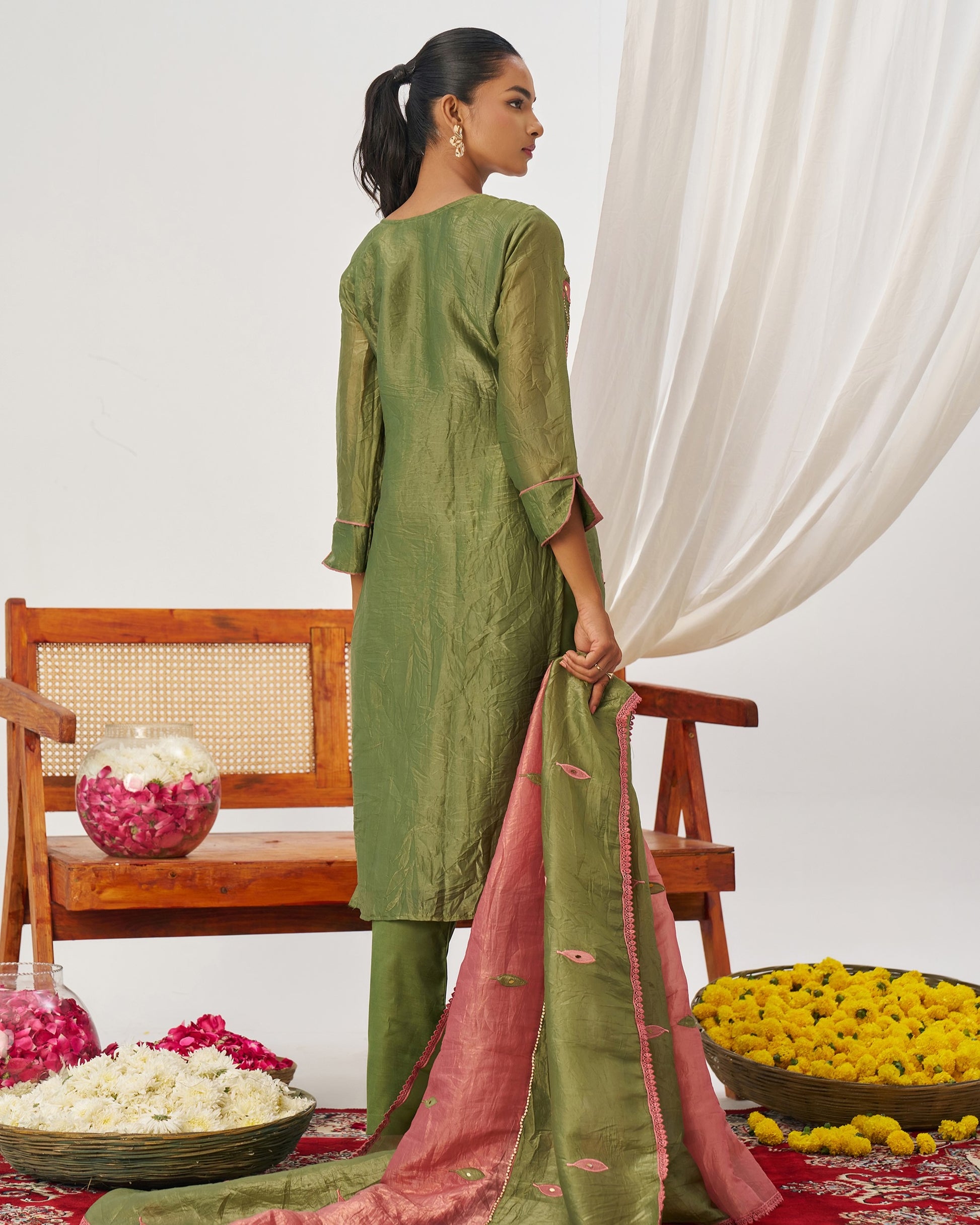 Woman in a green traditional outfit with a pink dupatta, standing in a decorated room with flowers and a chair.
