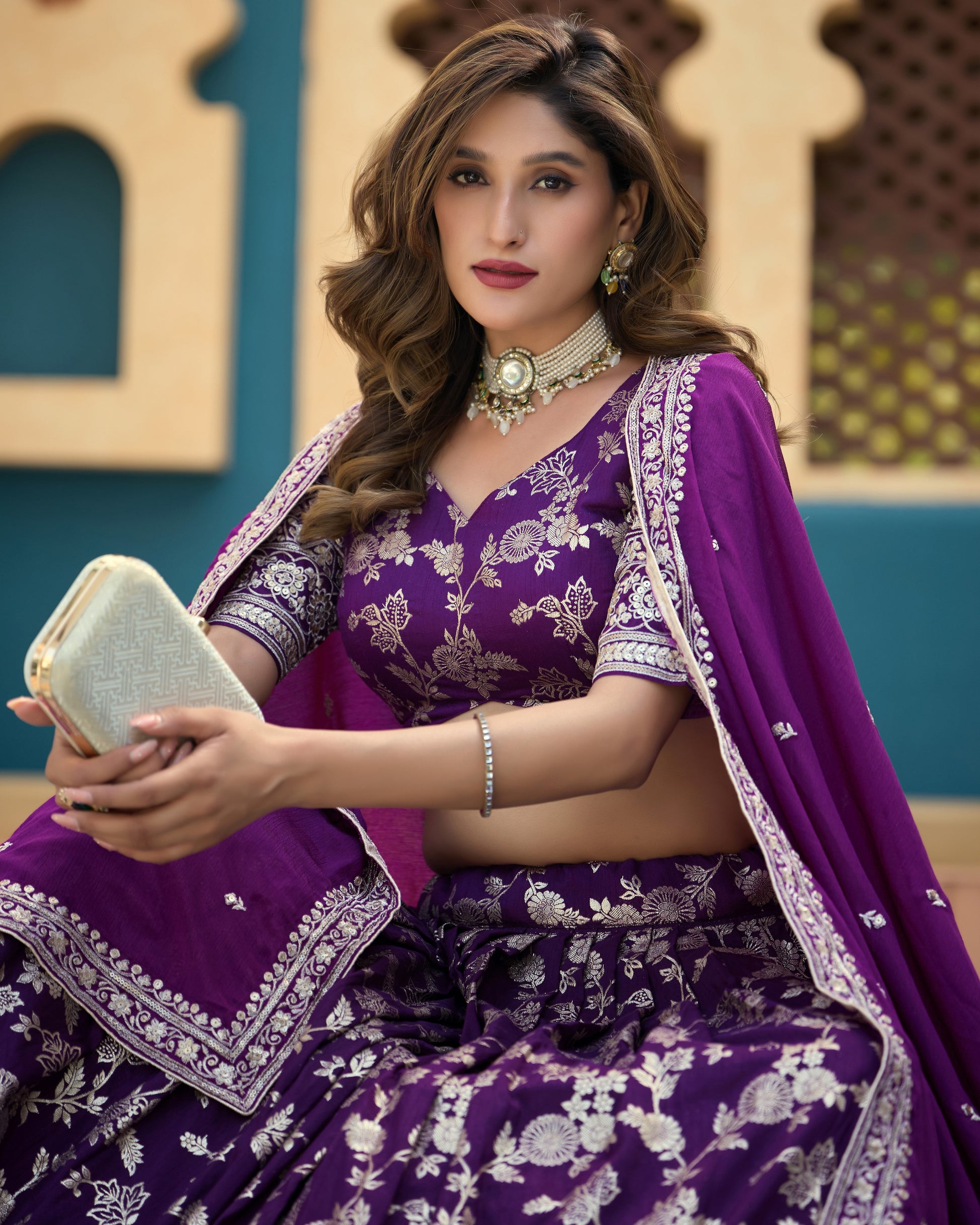Woman in a purple traditional outfit with white patterns, holding a clutch, against a decorative background.