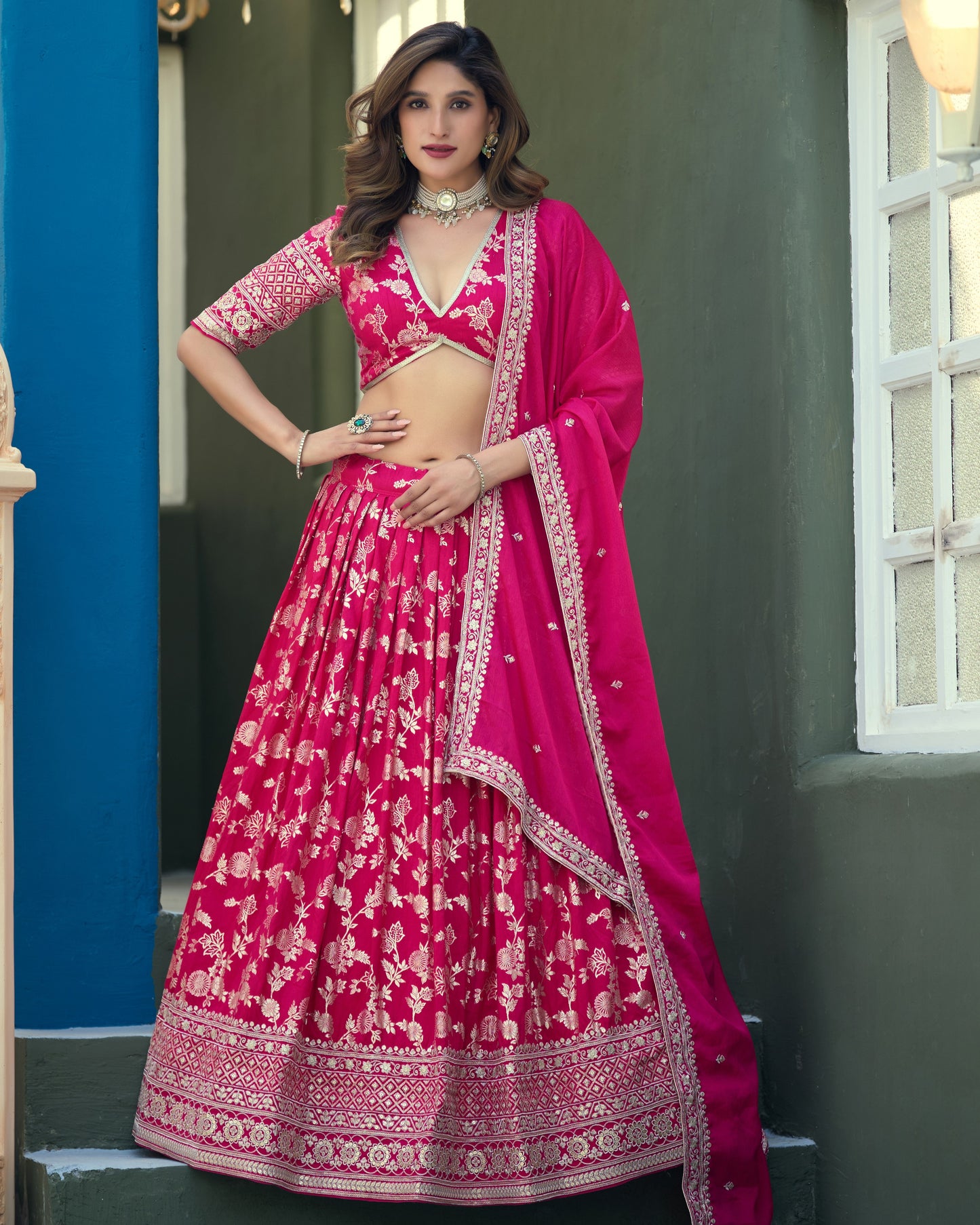 Woman in a pink and red traditional outfit standing against a blue and green wall.
