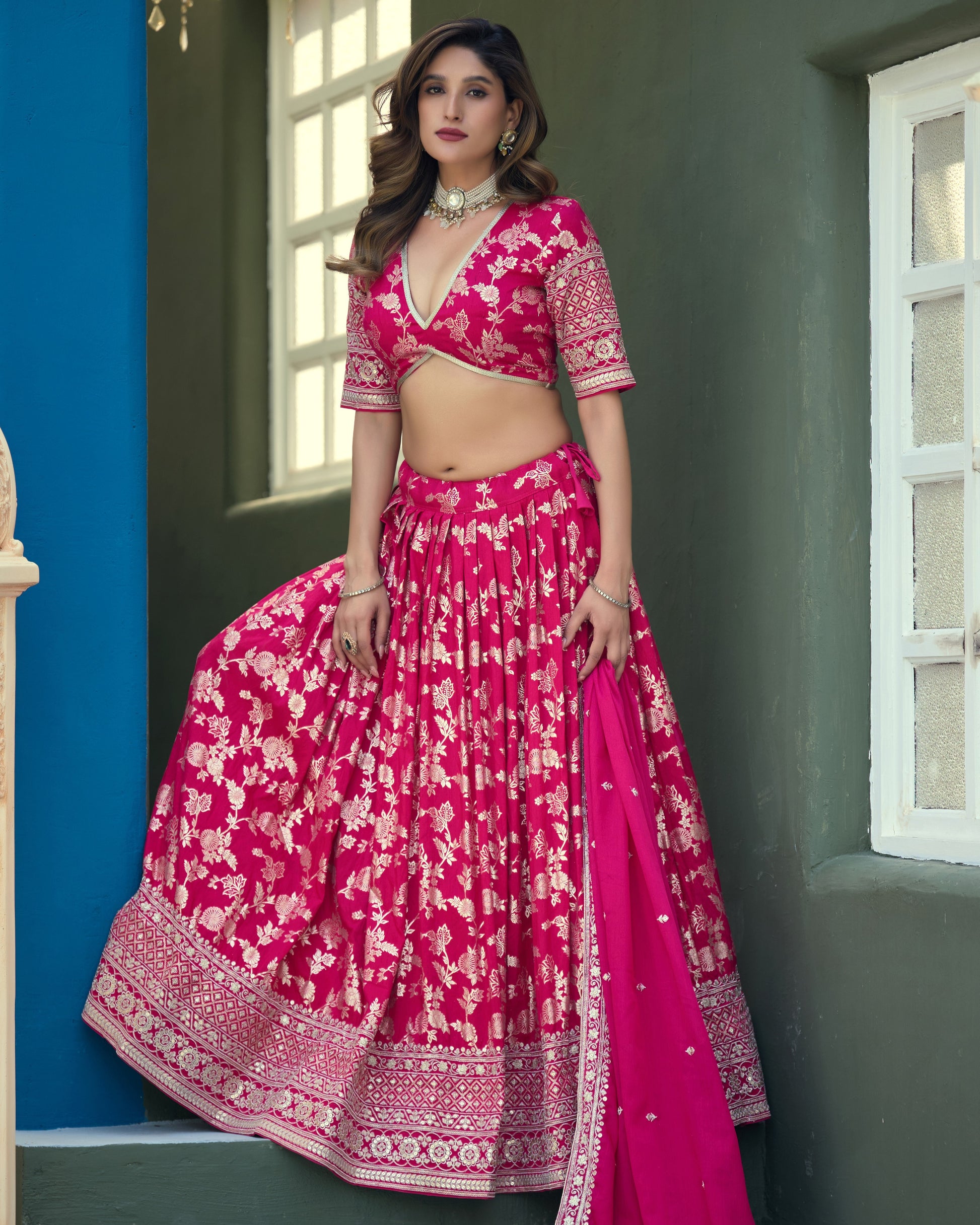 Woman wearing a pink traditional outfit with white patterns against a blue wall.