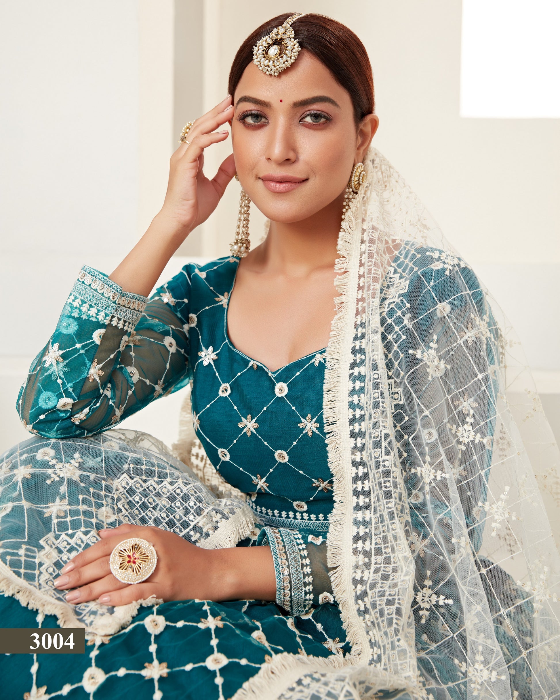Woman in a traditional teal and white outfit with jewelry, sitting indoors.