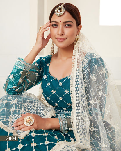 Woman in a traditional teal and white outfit with jewelry, sitting indoors.