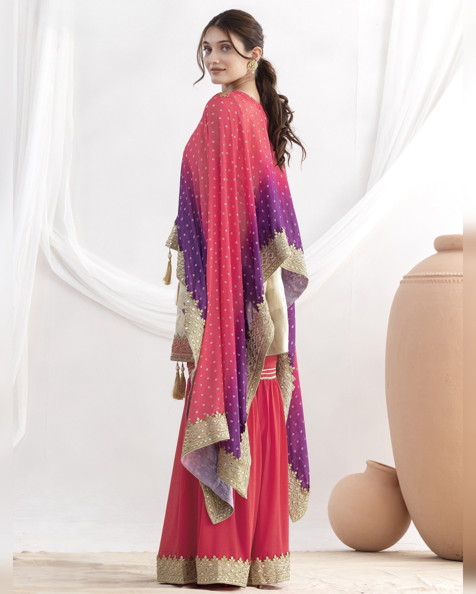 Woman wearing a pink and purple traditional outfit with a matching dupatta in a minimalistic setting.