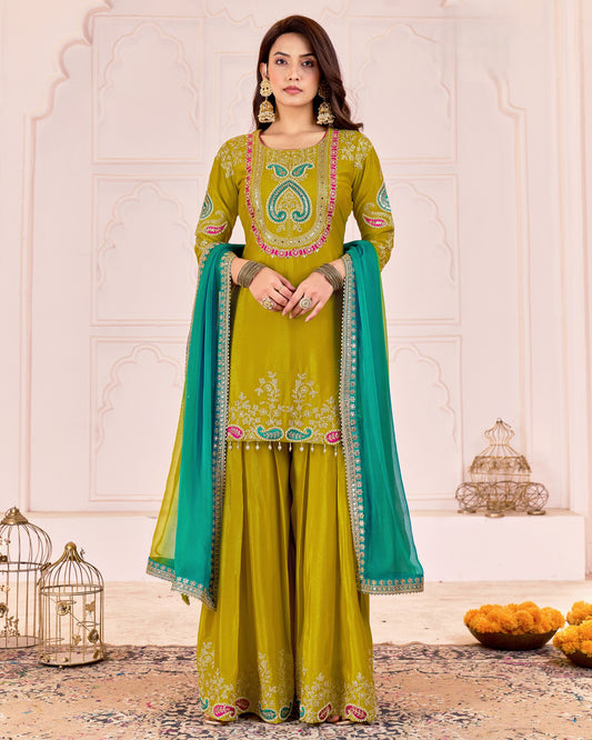 Woman wearing a green embroidered traditional outfit with a blue dupatta in a decorative setting.