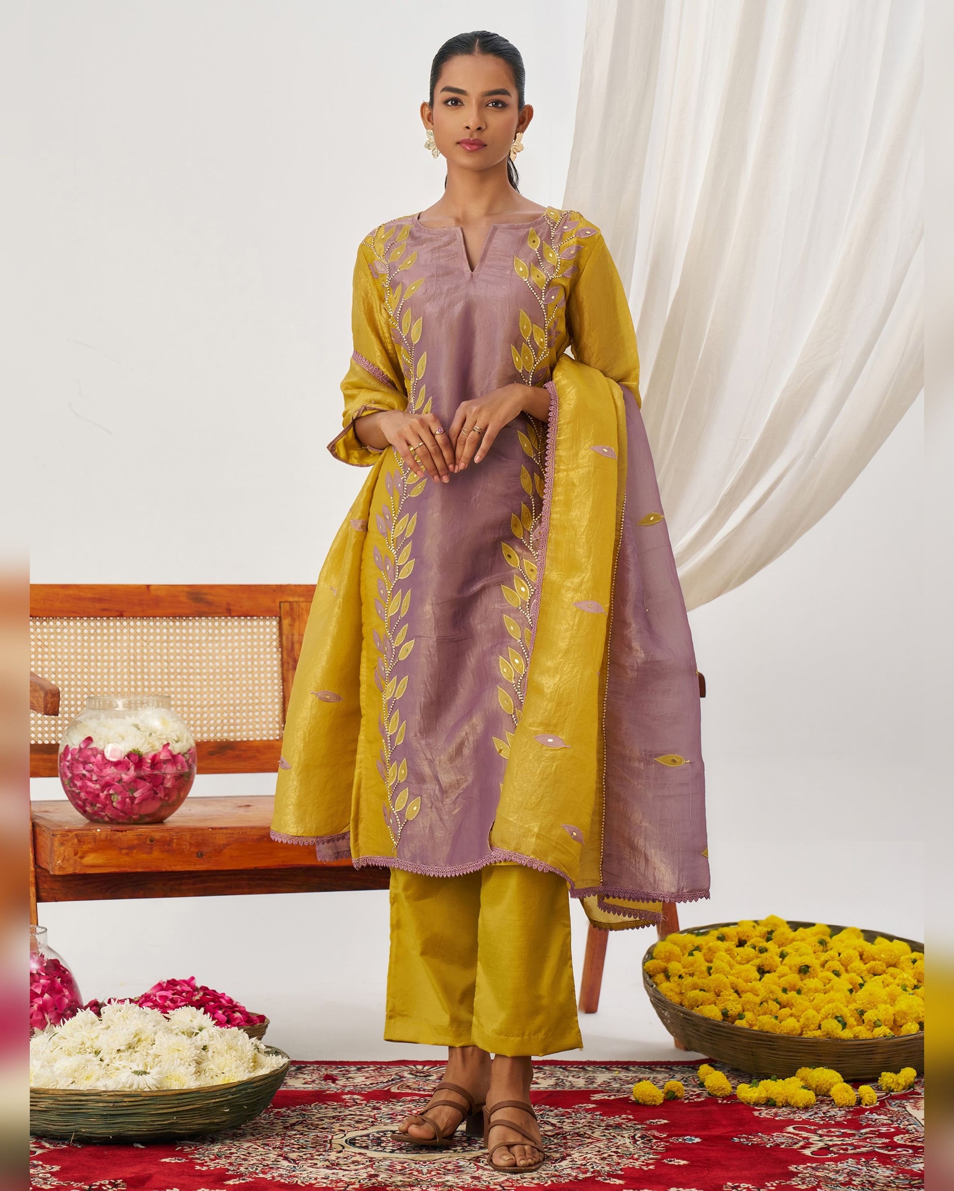 Woman in a yellow and purple traditional outfit standing in a decorated room with flowers and a bench.