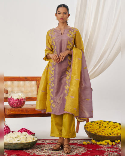 Woman in a yellow and purple traditional outfit standing in a decorated room with flowers and a bench.