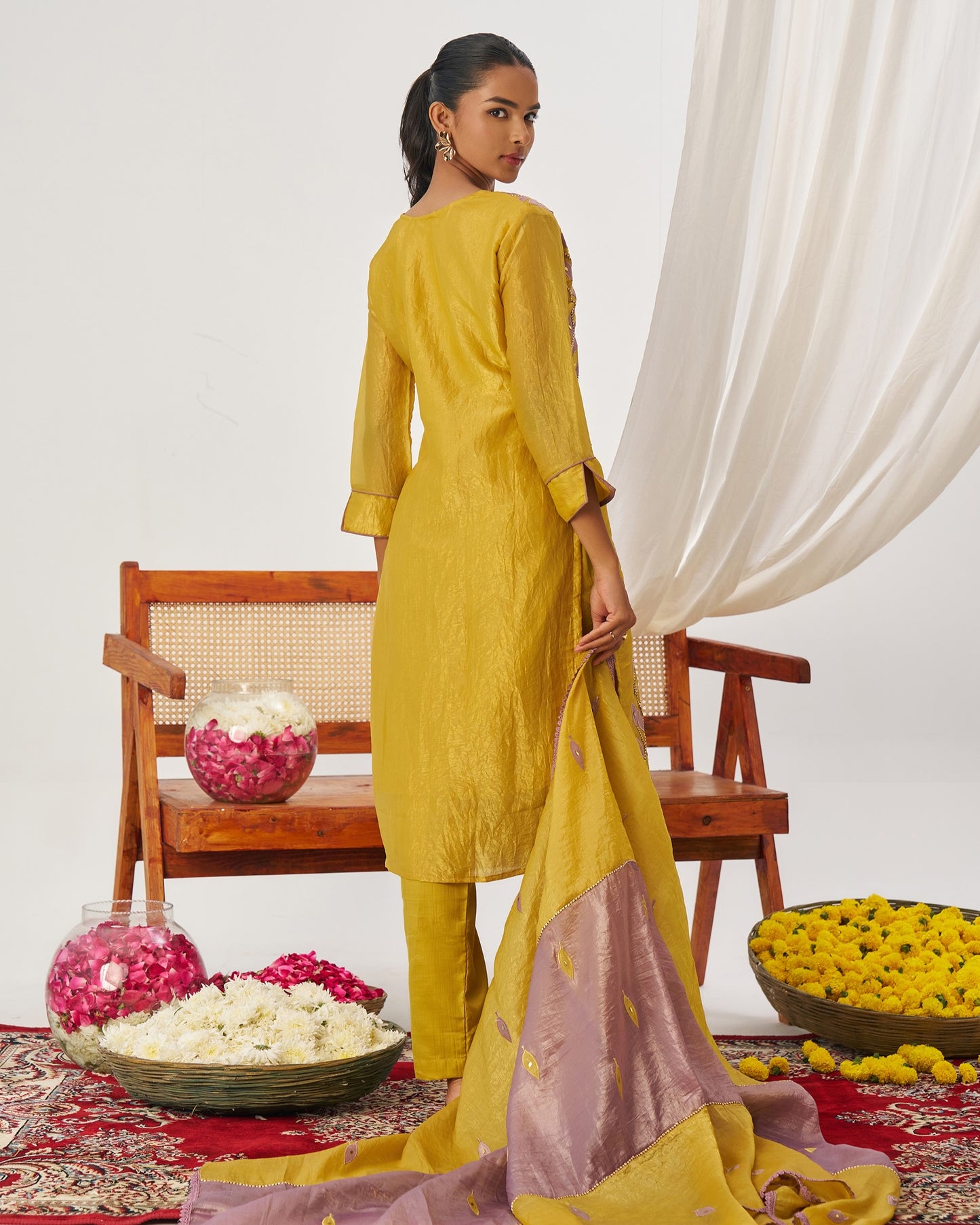 Woman in a yellow traditional outfit standing in a decorated room with flowers and a chair.