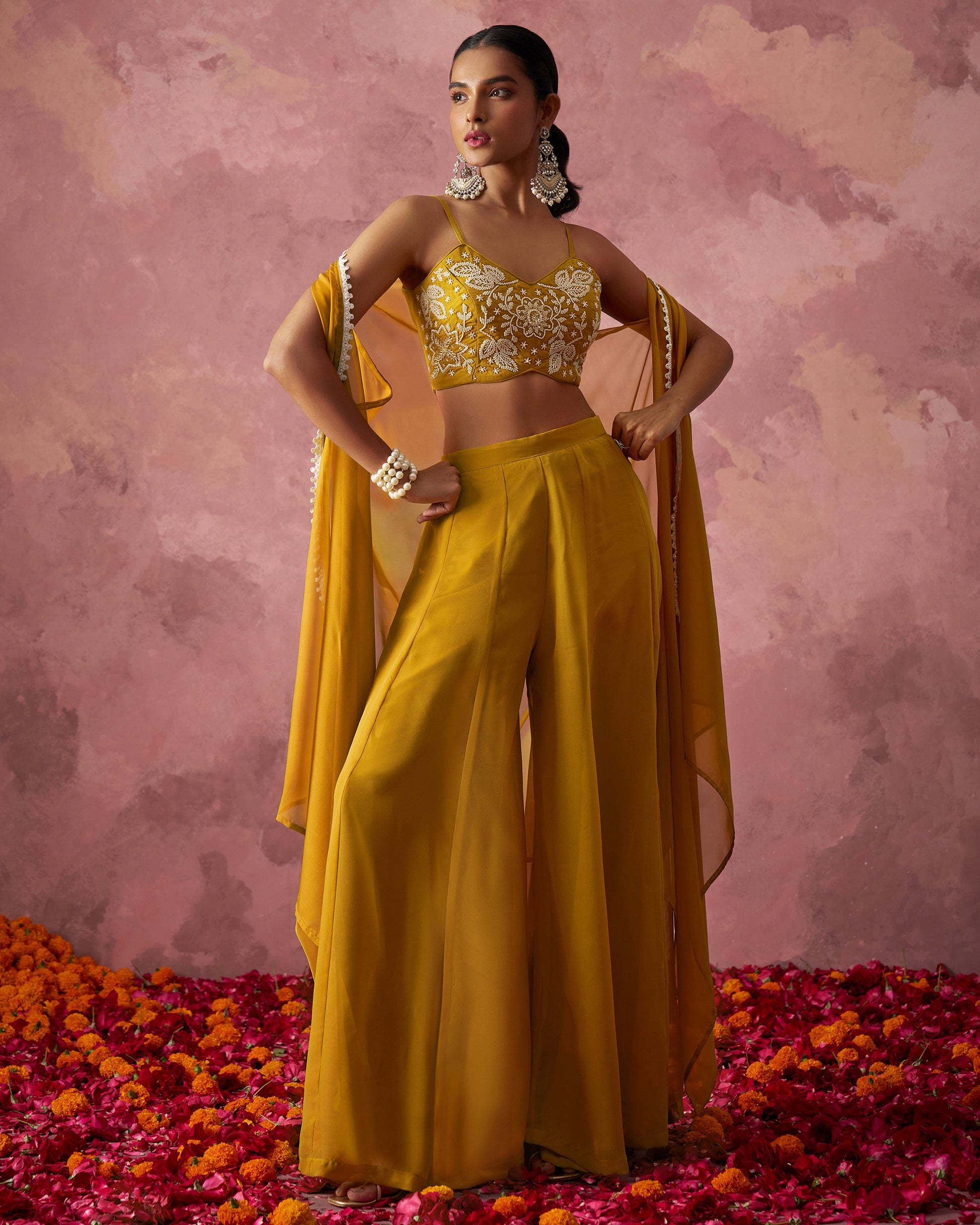 Woman in a yellow traditional outfit with a gold top and flowing skirt, standing on rose petals against a pink background.