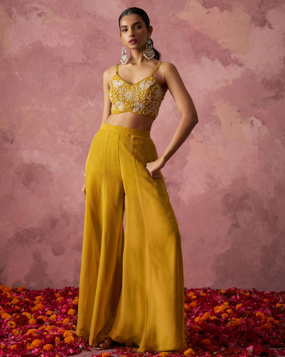 Woman in a yellow outfit with floral top and wide-leg pants standing on red and orange flower petals against a pink textured background.