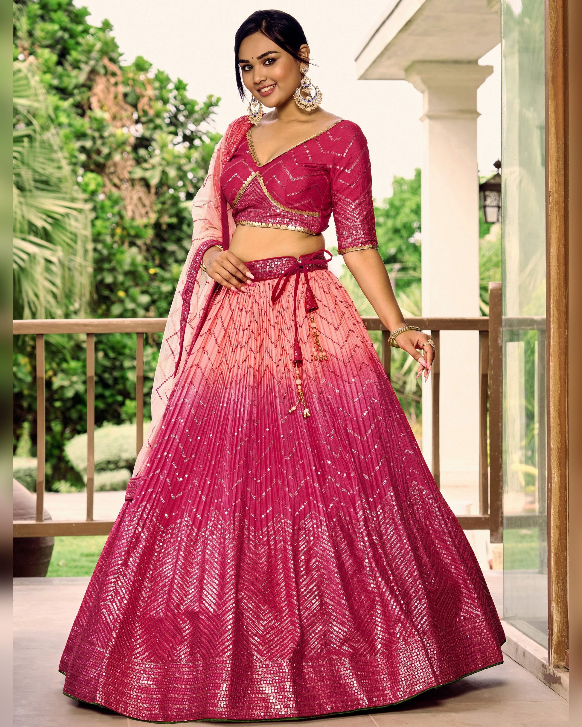 Woman wearing a pink and red lehenga with a gradient effect, standing on a balcony.