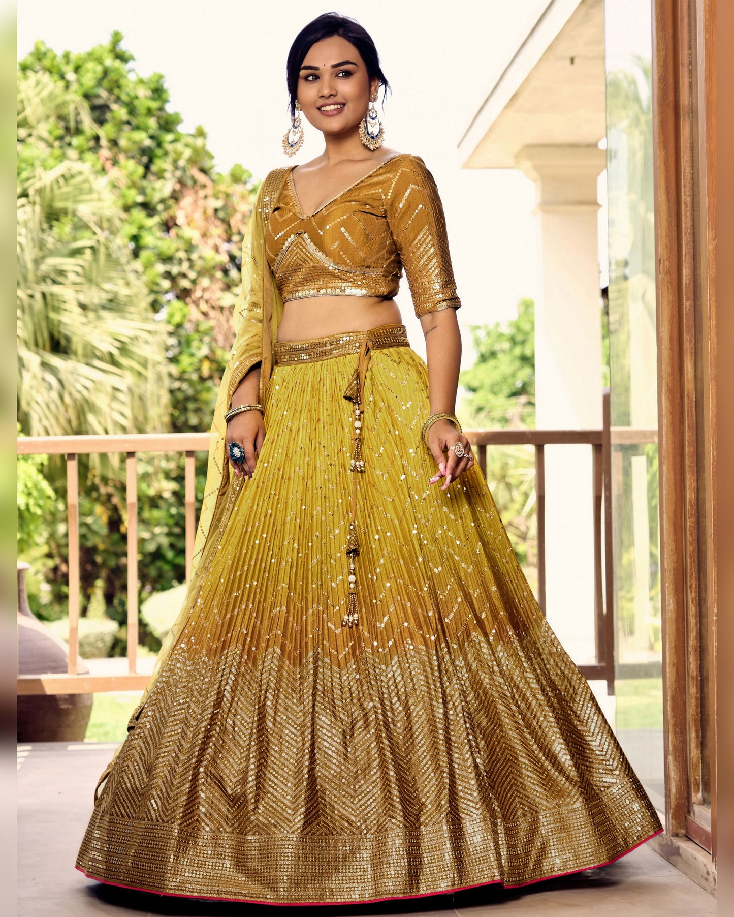 Woman wearing a traditional yellow and gold outfit on a balcony with greenery in the background