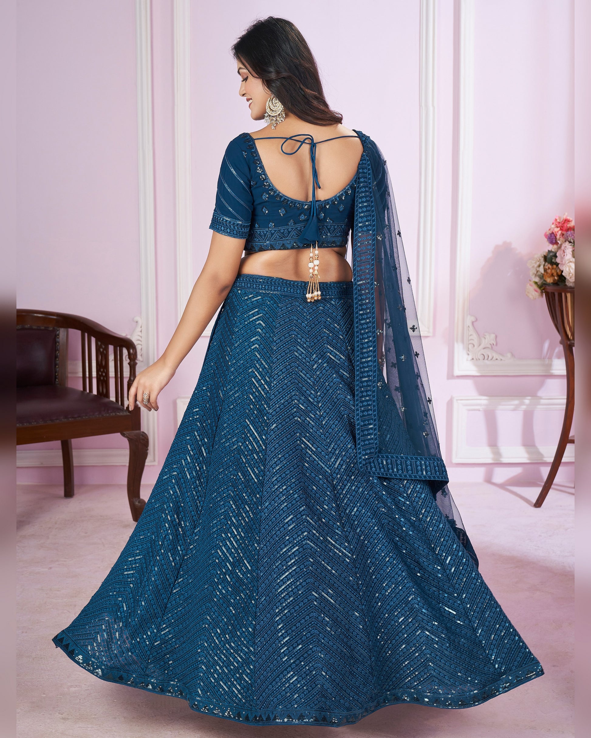 Woman wearing a blue traditional outfit in a room with pink walls and a wooden chair.