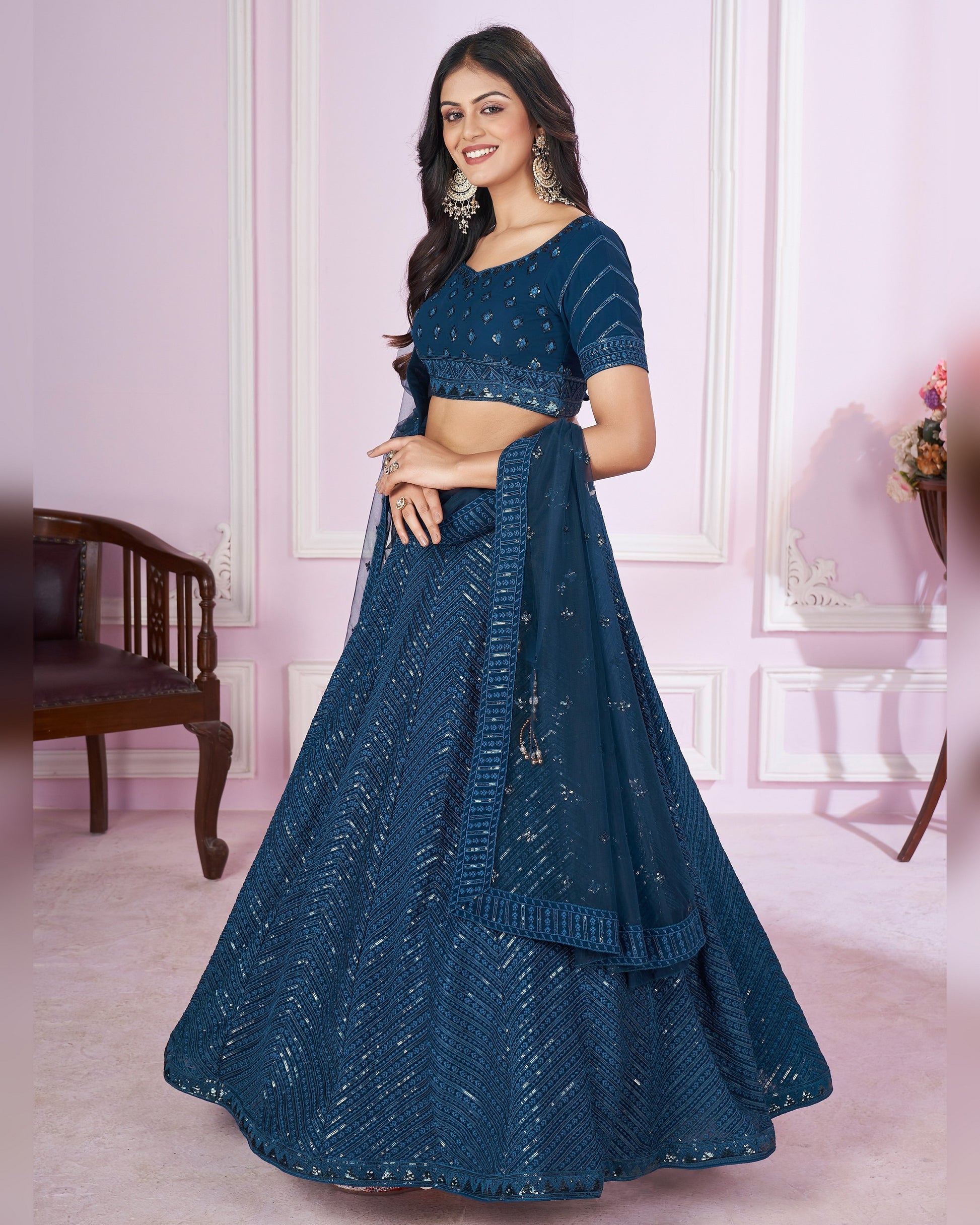 Woman wearing a blue embroidered lehenga in a room with pink walls and wooden furniture.