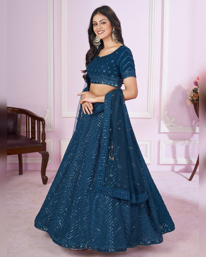 Woman wearing a blue embroidered lehenga in a room with pink walls and wooden furniture.