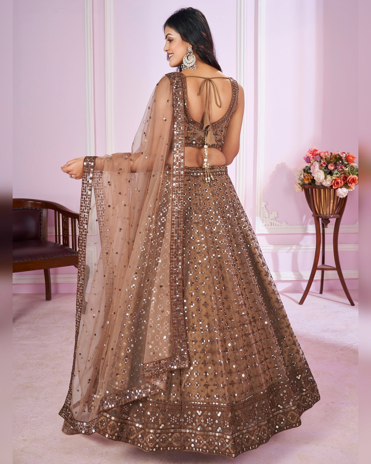 Woman wearing a brown embroidered lehenga in a room with pink walls and a basket of flowers.