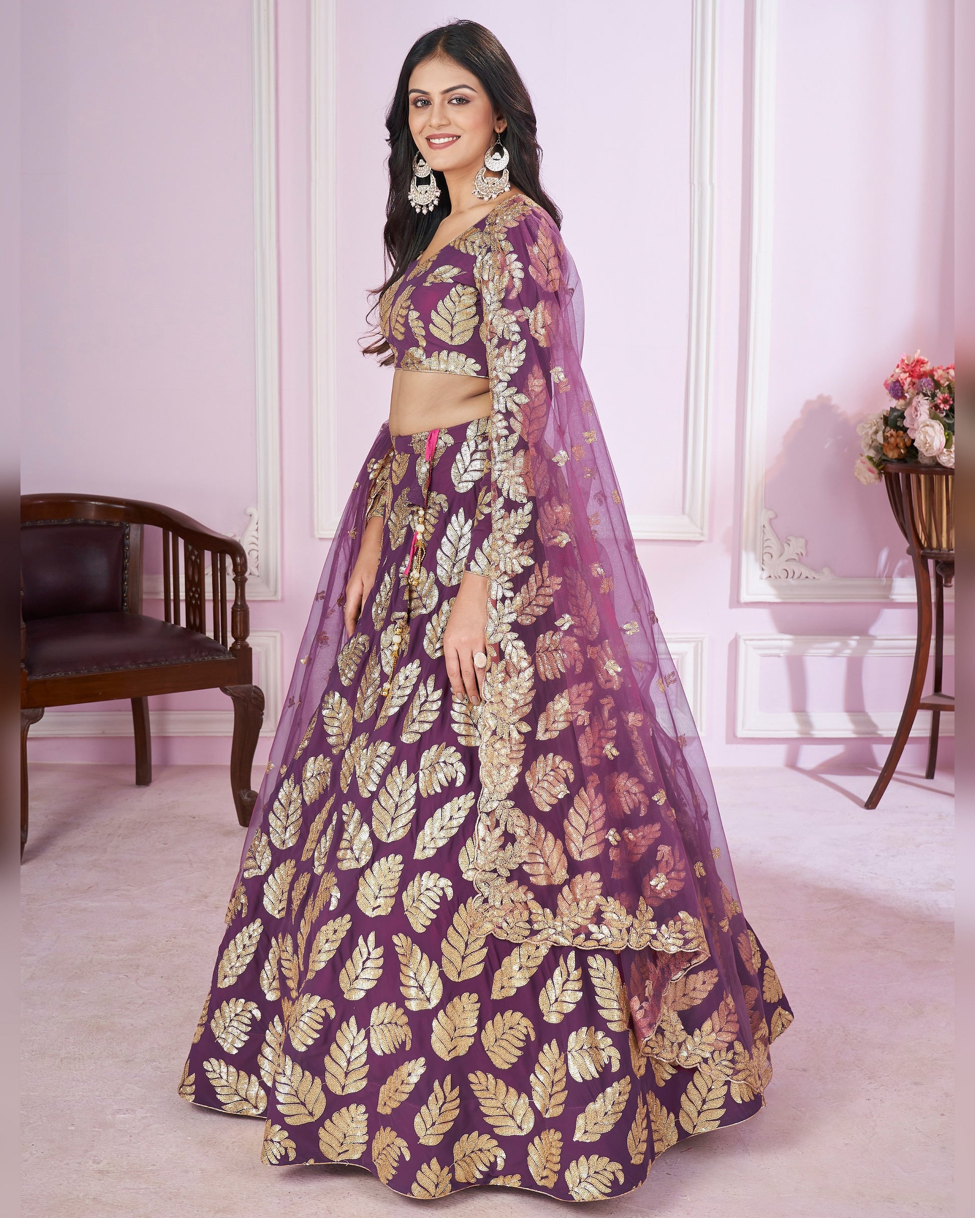 Woman wearing a purple and gold traditional outfit in a room with white walls and a wooden chair.
