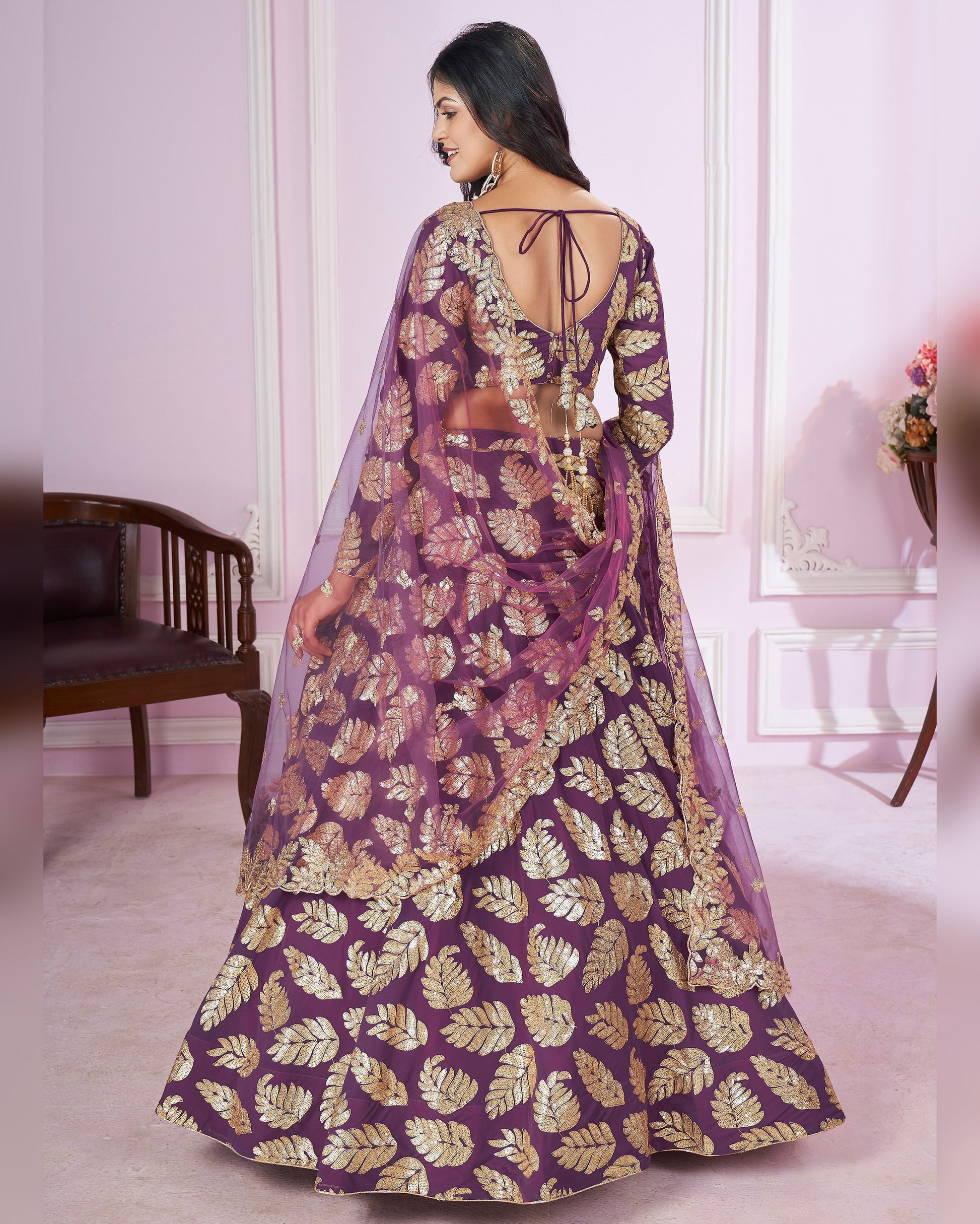 Woman wearing a purple and gold patterned saree in a room with a chair and table.