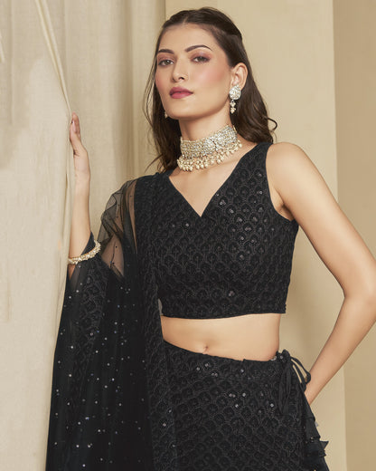 Woman wearing a black embroidered outfit with jewelry against a beige curtain background