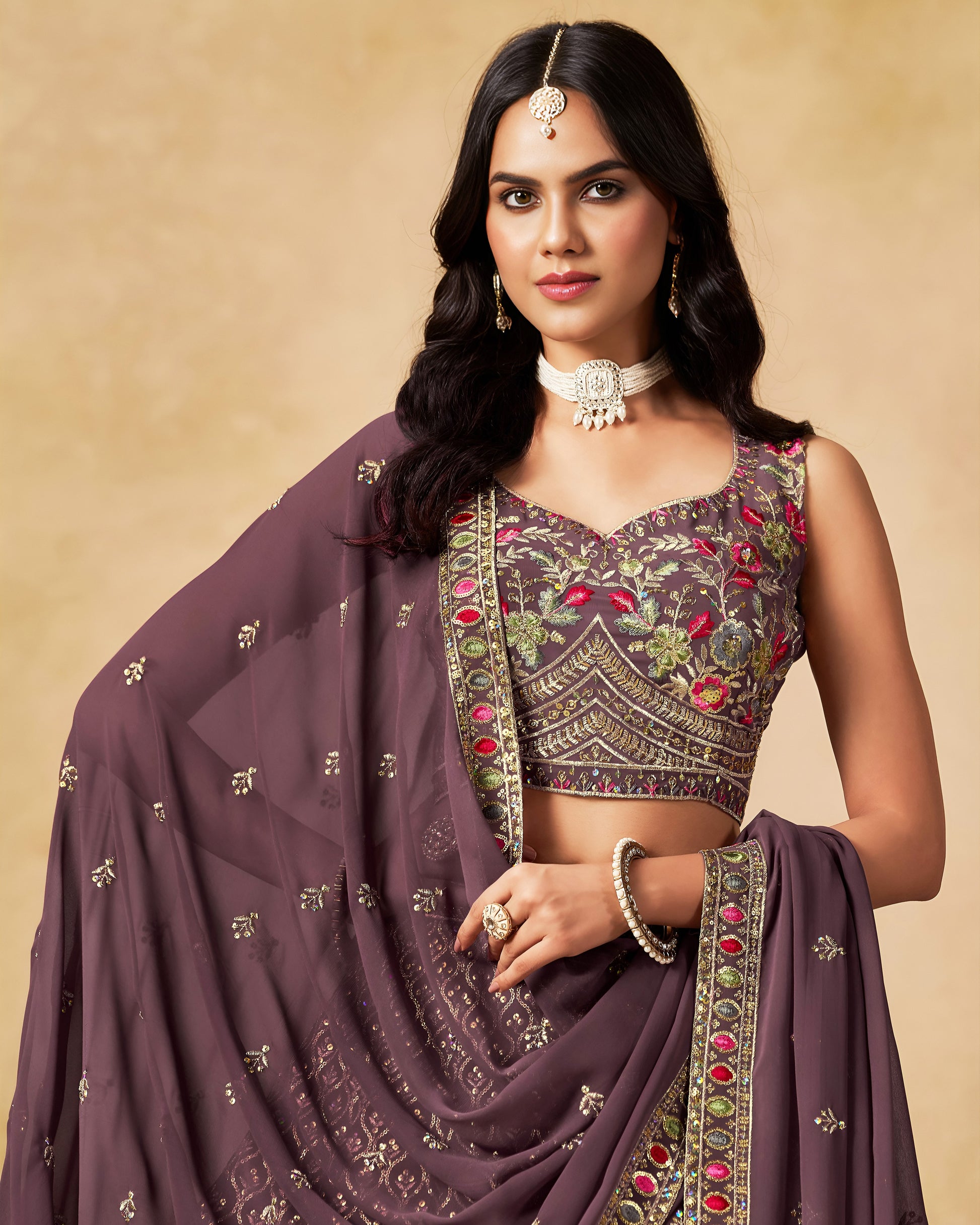 Woman wearing a traditional outfit with a purple saree and embroidered blouse against a beige background