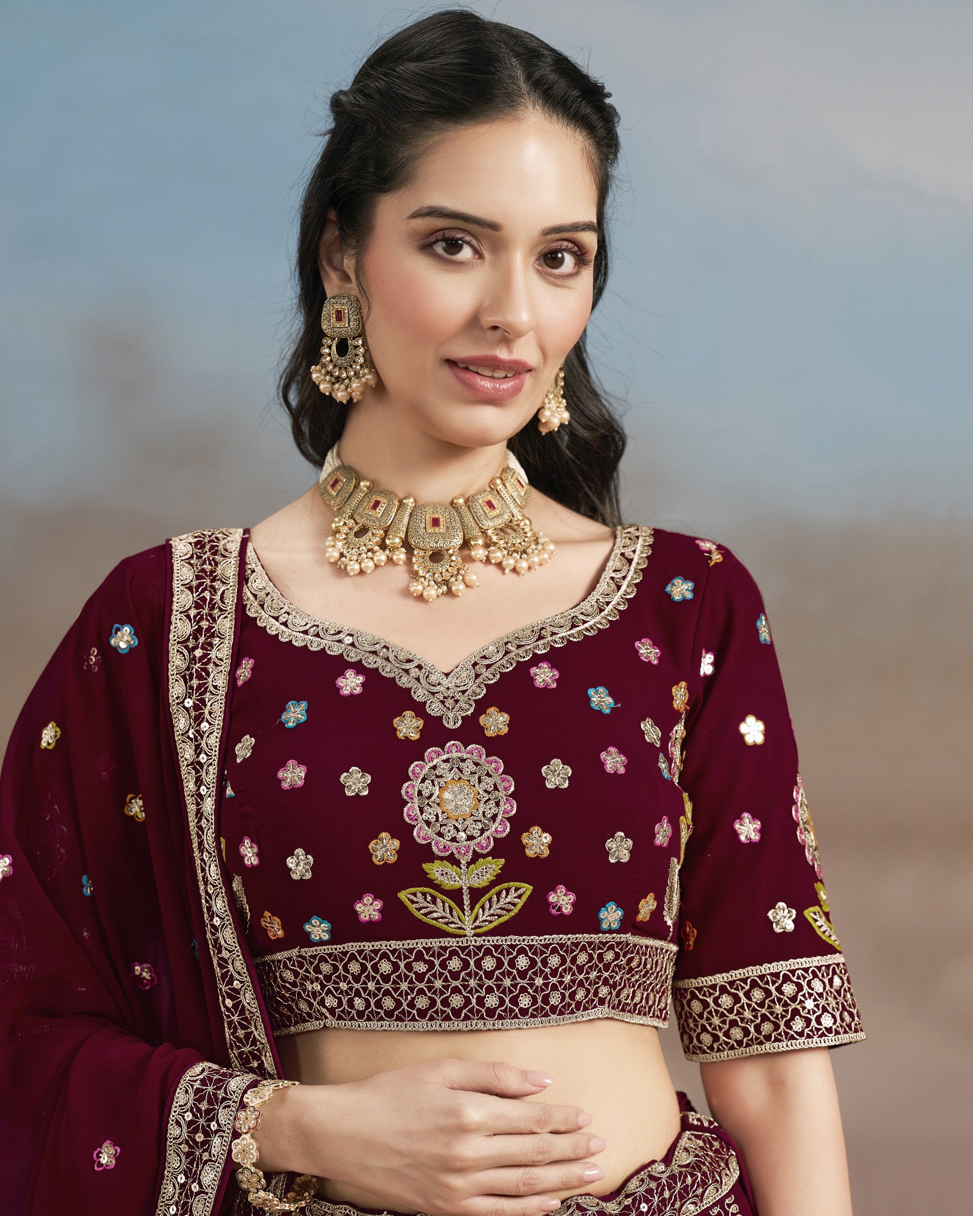 Woman wearing a maroon traditional outfit with floral patterns and gold jewelry against a blurred background