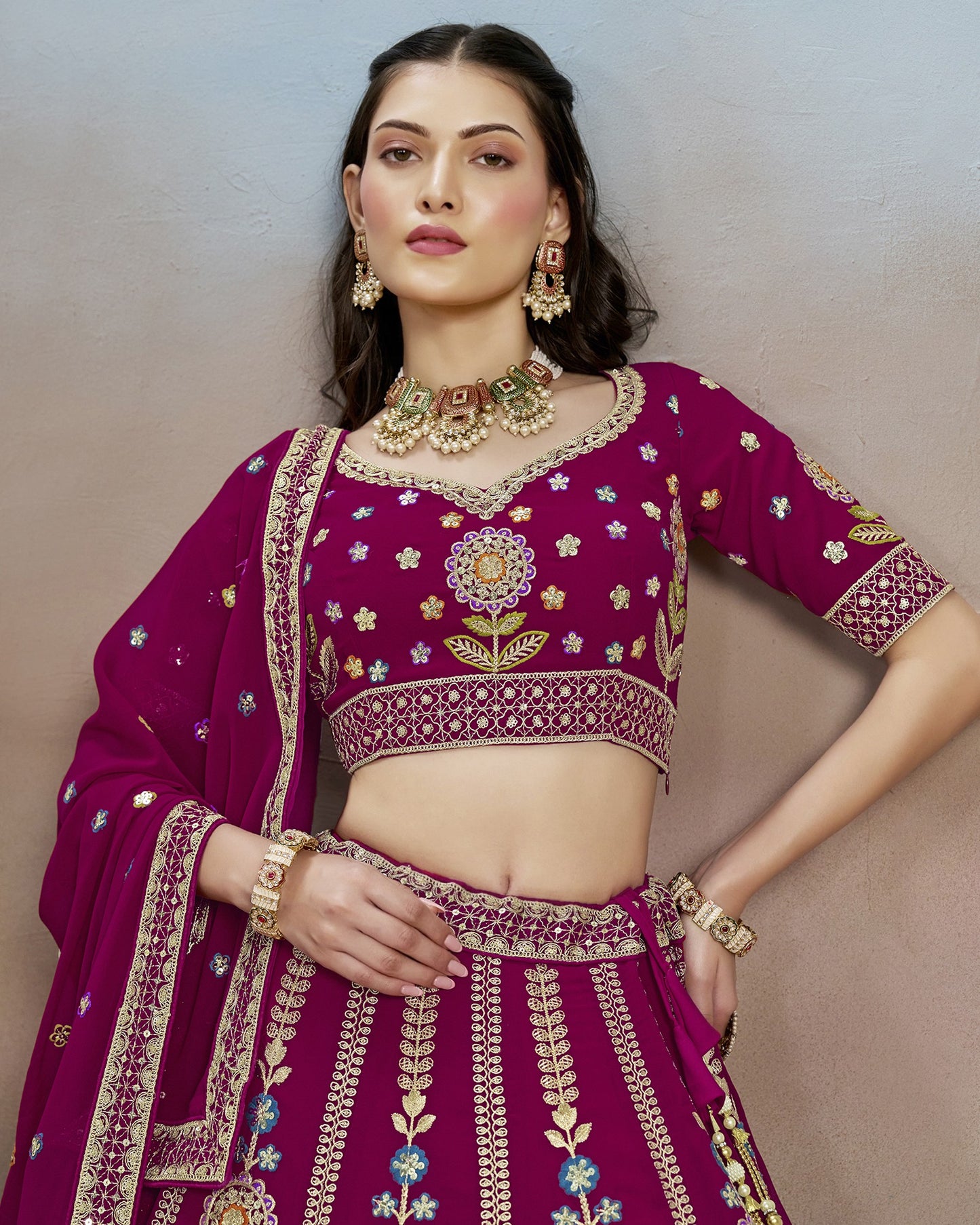 Woman wearing a traditional purple embroidered outfit with gold jewelry against a neutral background