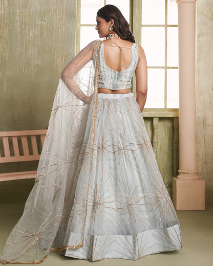Woman in a silver embroidered lehenga standing in a room with a window and bench.