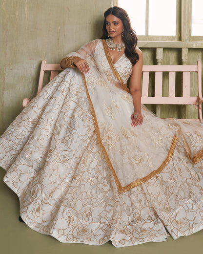 Woman in a white and gold embroidered traditional outfit sitting on a wooden chair.