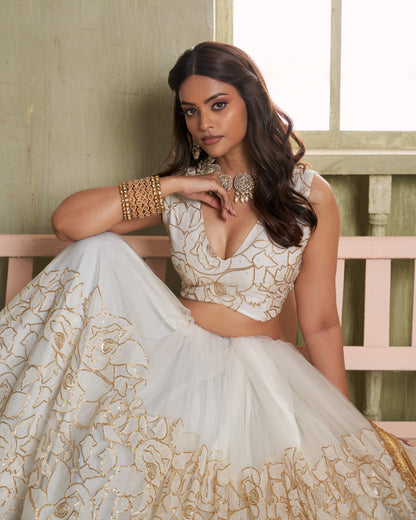 Woman in a white and gold embroidered traditional outfit sitting on a wooden bench.