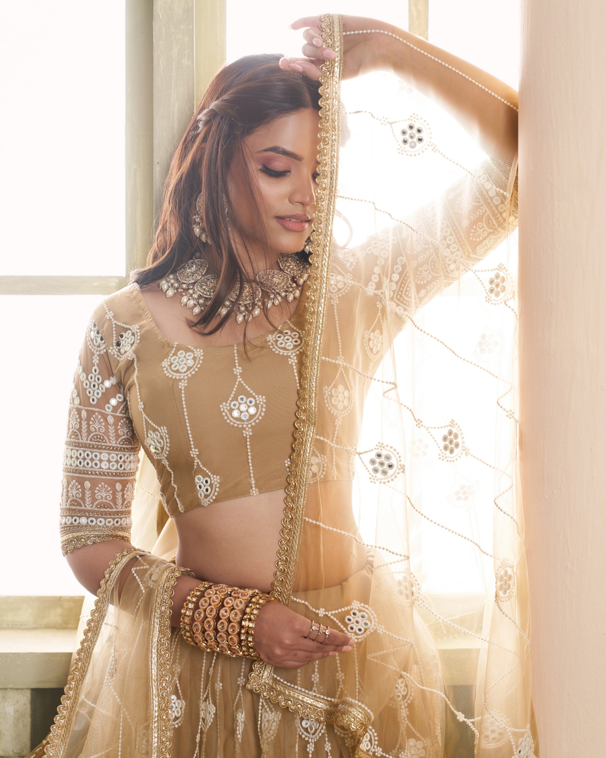 Woman in traditional beige outfit with intricate patterns and jewelry, standing indoors.