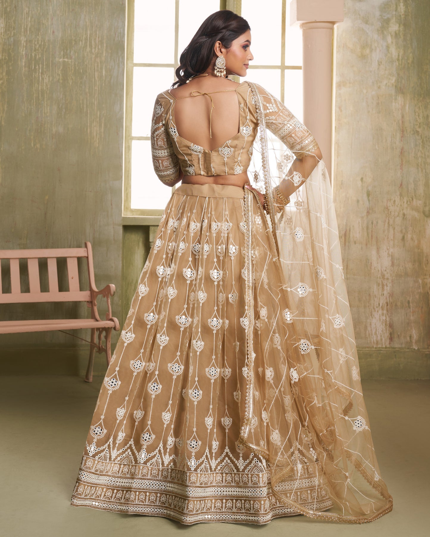 Woman wearing a beige embroidered traditional outfit in a room with a window and bench.
