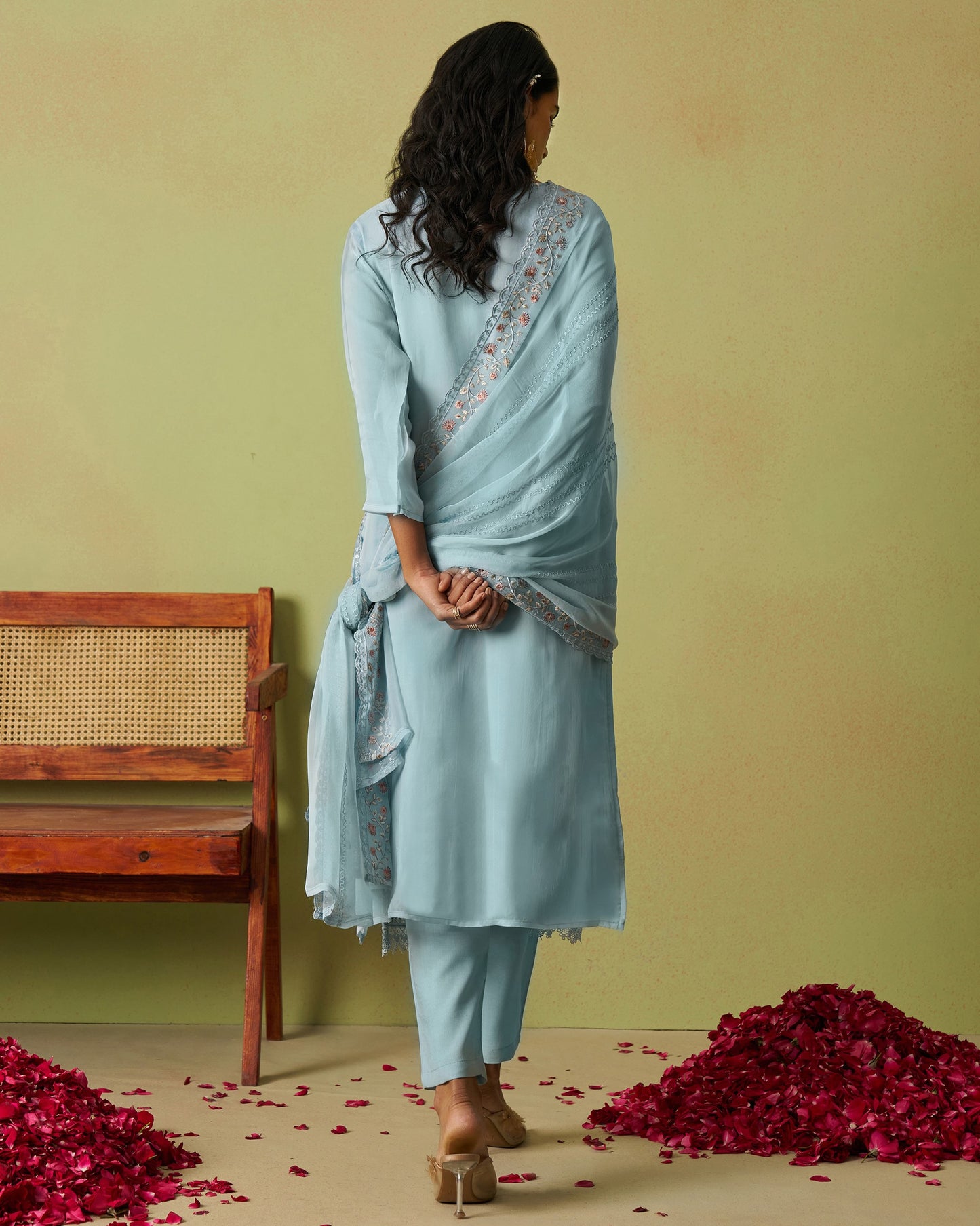 Woman in a light blue traditional outfit with a saree, standing against a green wall with red flower petals on the floor.