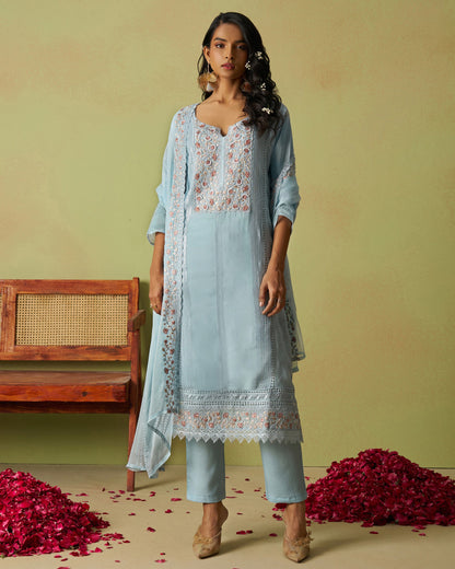 Woman wearing a light blue traditional outfit with floral patterns in a decorative setting with red flowers and a wooden bench.