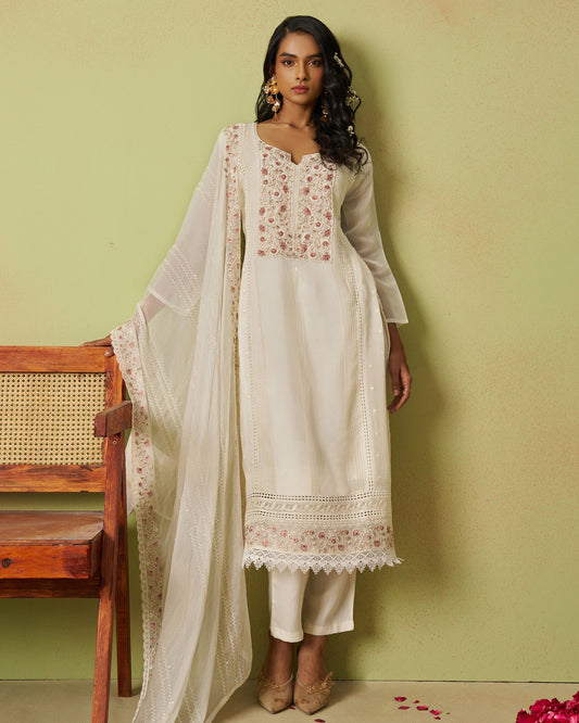 Woman wearing a white embroidered outfit with a sheer dupatta against a green wall.