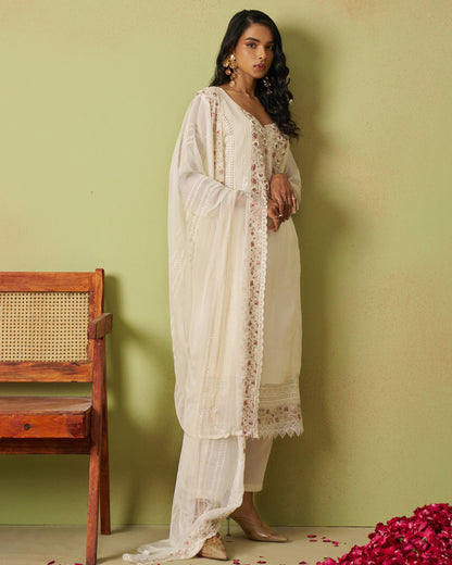 Woman in a white traditional outfit standing against a green wall with a wooden chair and red flowers on the floor.