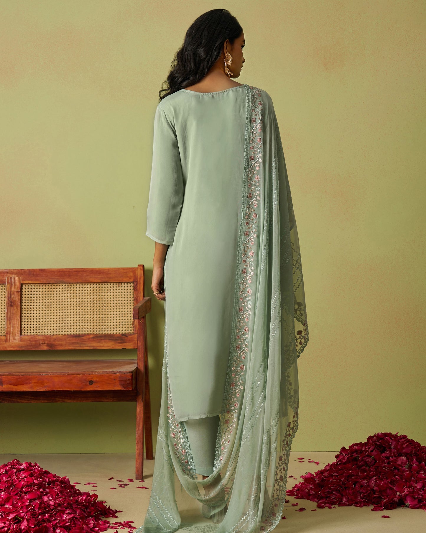 Woman wearing a light green outfit with a matching dupatta in a room with a wooden bench and red flowers.