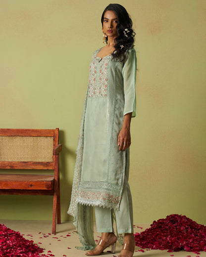 Woman in a light green traditional outfit with floral embroidery standing against a plain background.