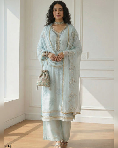 Woman wearing a light blue embroidered traditional outfit in a bright room.