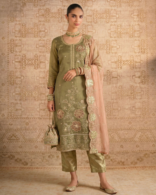 Woman wearing a traditional green embroidered outfit with a pink dupatta against a patterned wall.