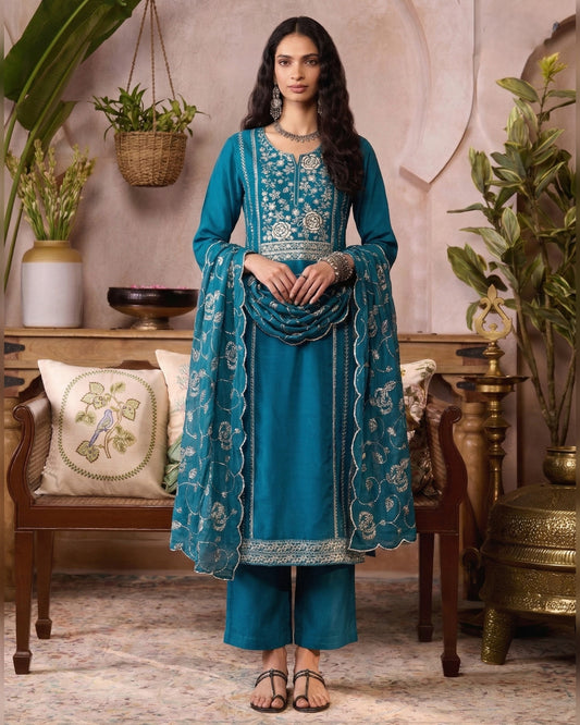 Woman in a blue embroidered traditional outfit standing in an elegant room with decorative elements.