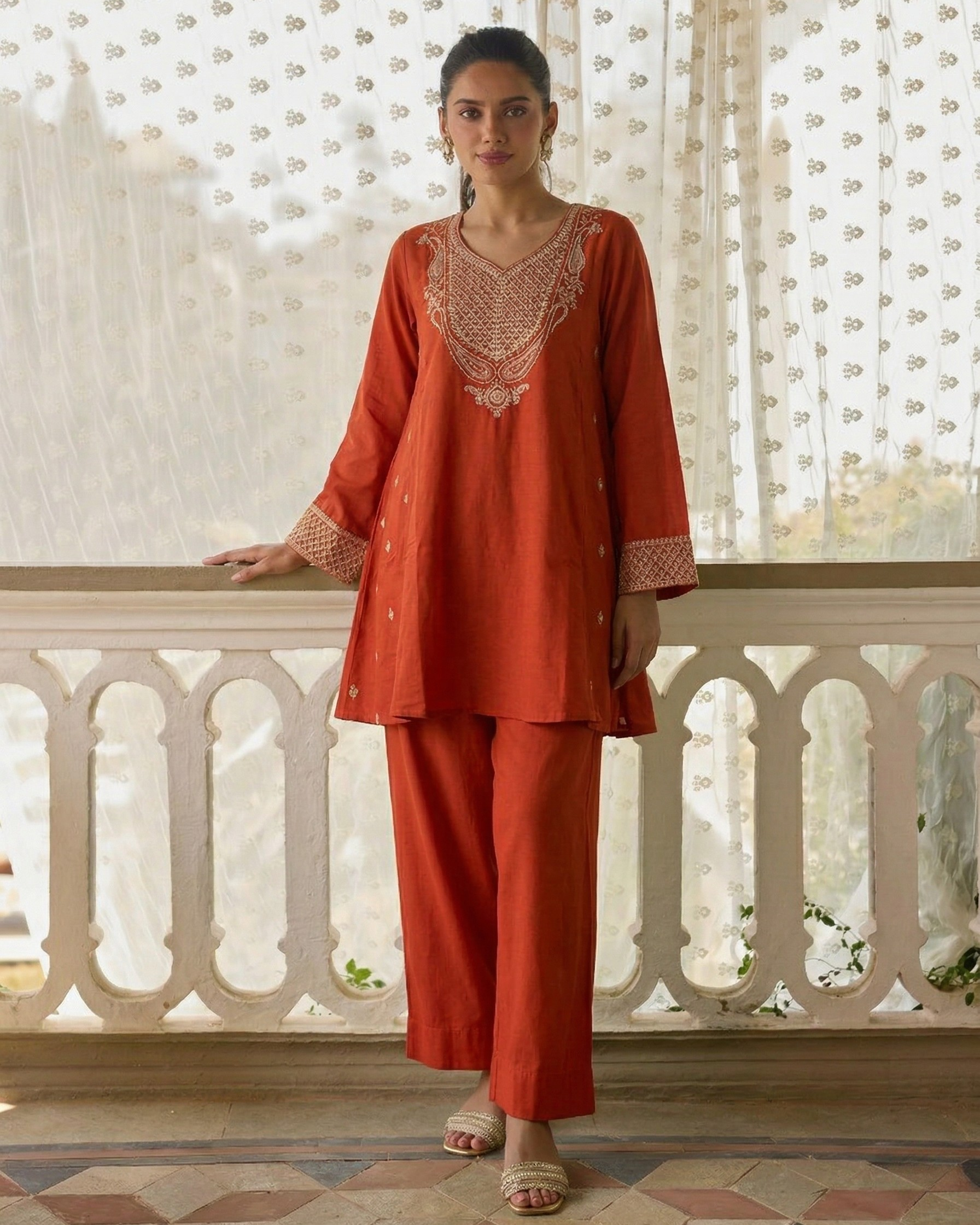 Woman wearing an orange traditional outfit with intricate patterns in a decorative interior setting.