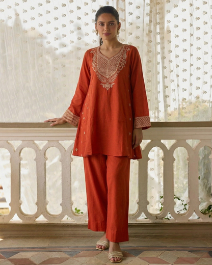 Woman wearing an orange traditional outfit with intricate patterns in a decorative interior setting.