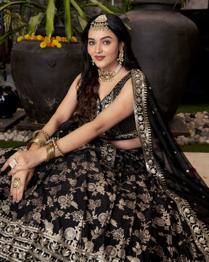 Woman in traditional black and gold outfit with jewelry sitting outdoors.