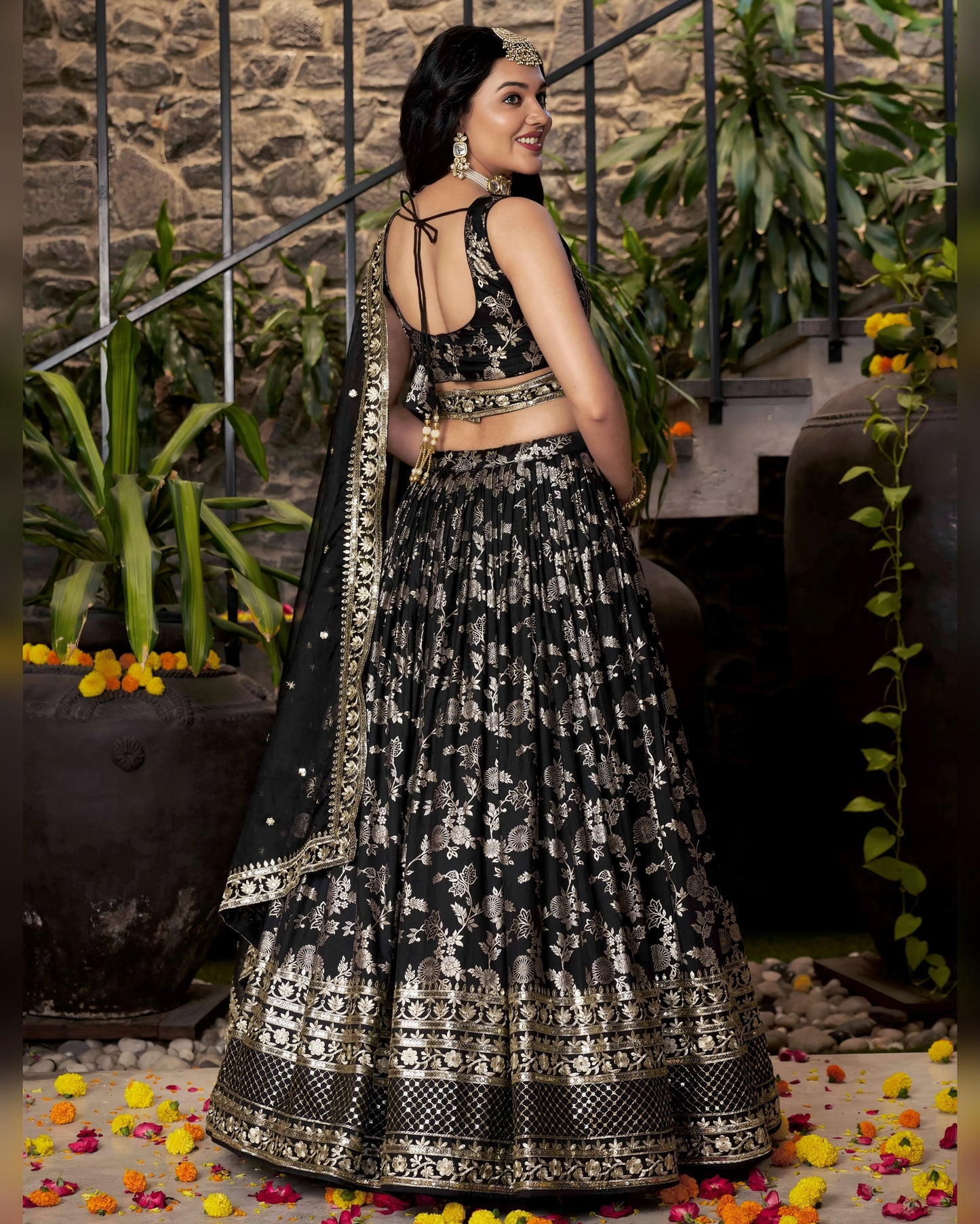 Woman in a black and white patterned traditional outfit standing in a decorative setting with flowers and plants.
