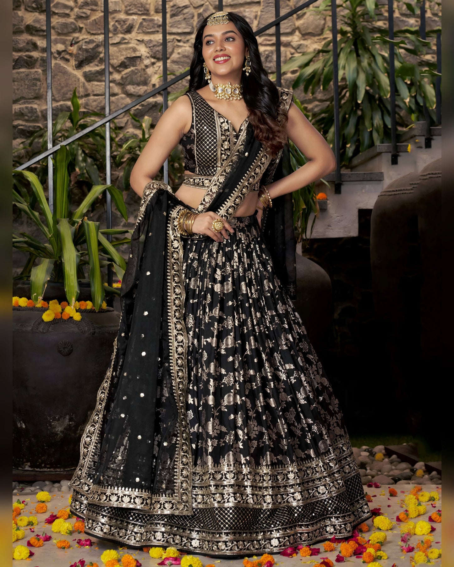 Woman in a black and silver traditional outfit standing in front of a stone wall with plants.