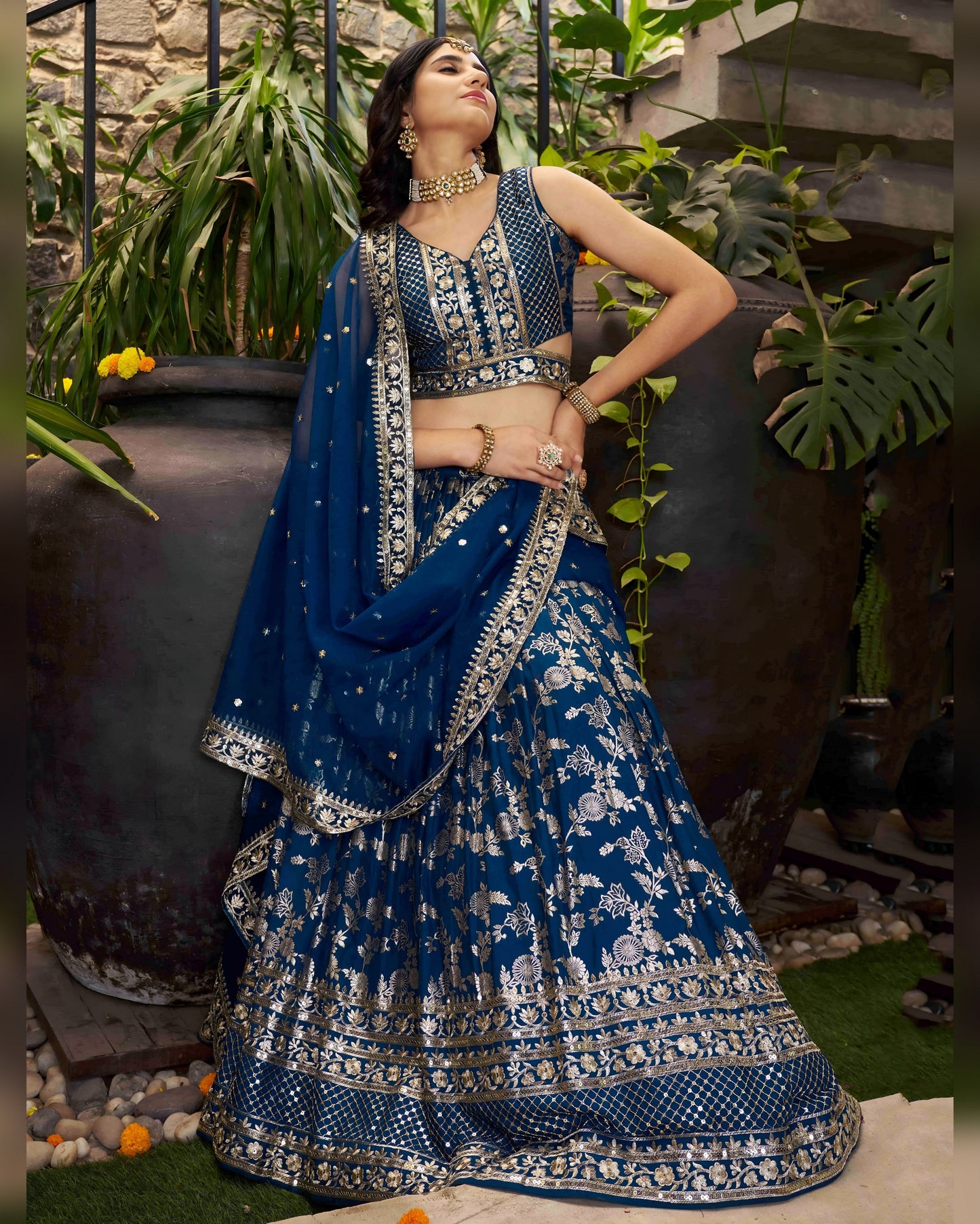 Woman in a blue and silver traditional outfit standing outdoors with plants in the background