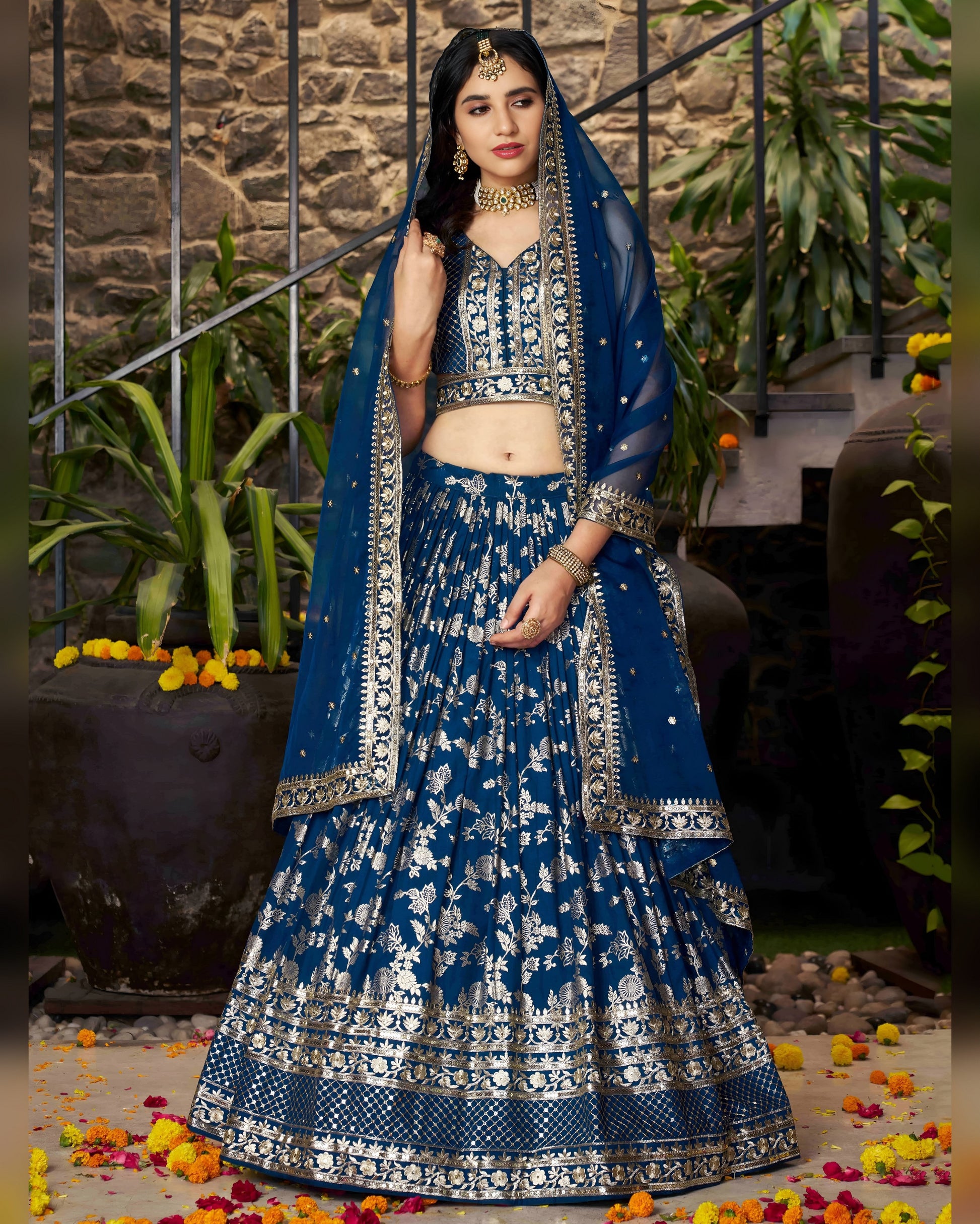 Woman in a blue and silver traditional outfit standing outdoors with plants and flowers around.