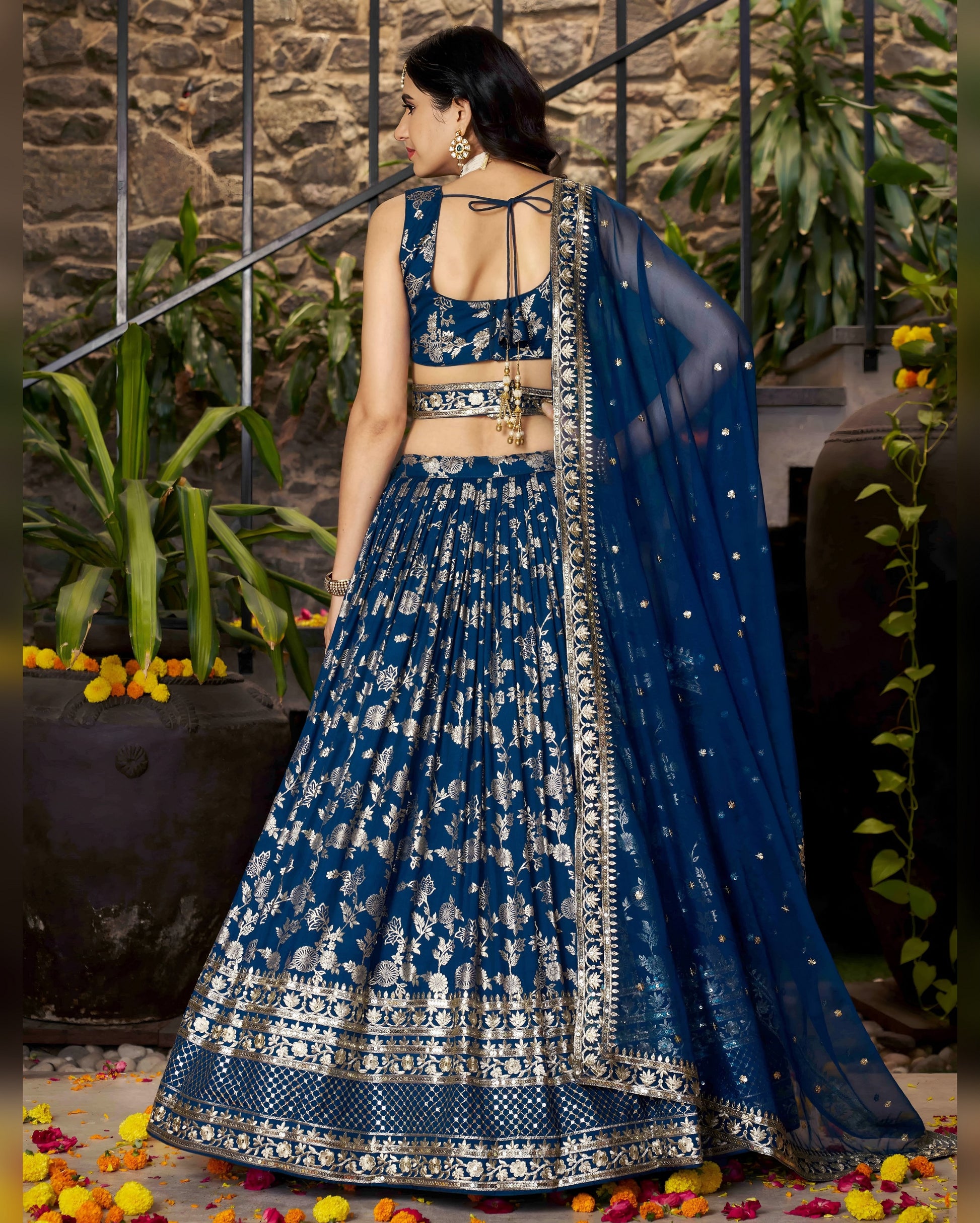Woman in a blue and white traditional outfit standing in an outdoor setting with plants and flowers.