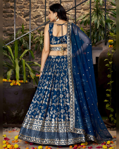 Woman in a blue and white traditional outfit standing in an outdoor setting with plants and flowers.